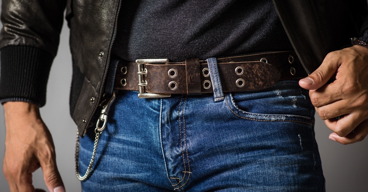 A man, shown from the waist up, posing in jeans, a dark leather belt, a black shirt, and a black leather jacket.