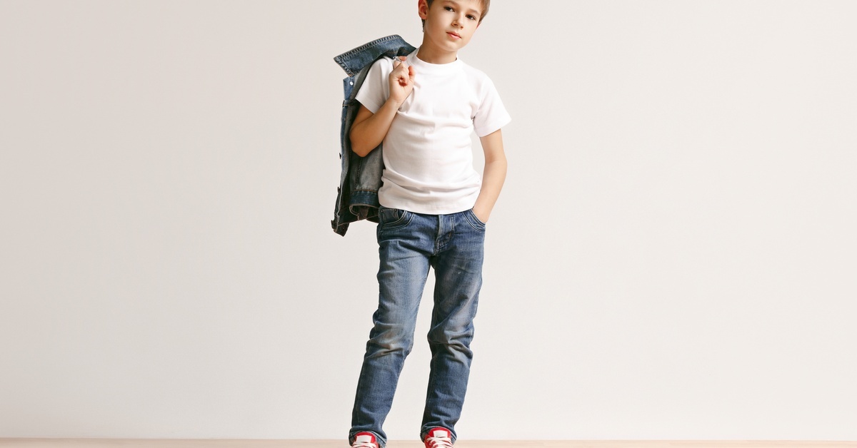 A young boy dressed in sneakers, jeans, and a white t-shirt posing, casually draping a denim jacket over his right shoulder.