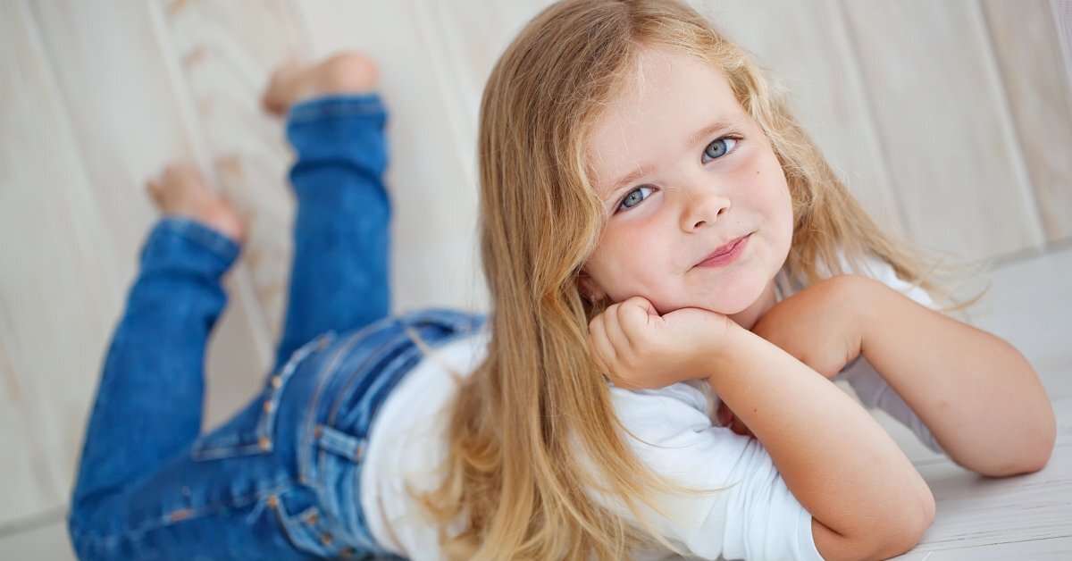A young girl with long blonde hair, wearing a white shirt and jeans, lying on her stomach with her face resting on her hands.