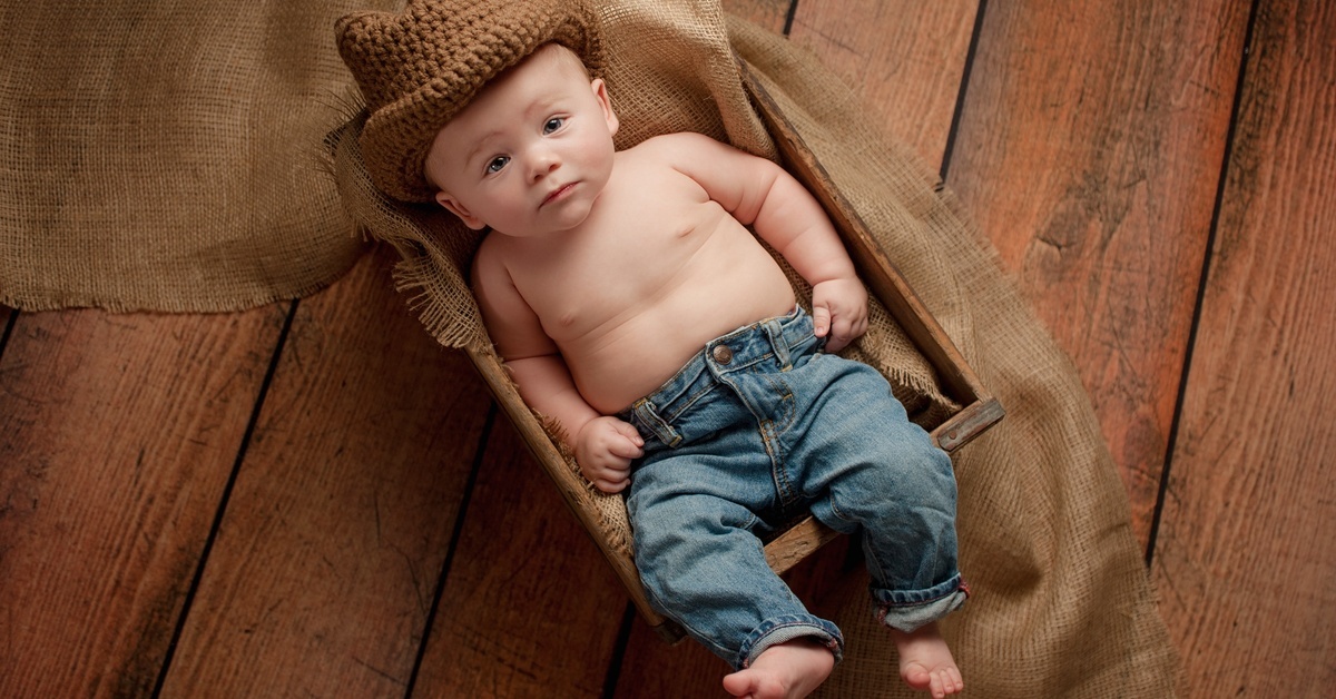 A baby in jeans and a crochet cowboy hat lying on a thin woven blanket in a bassinet on a wooden floor, looking up. A baby in jeans and a crochet cowboy hat lying on a thin woven blanket in a bassinet on a wooden floor, looking up.