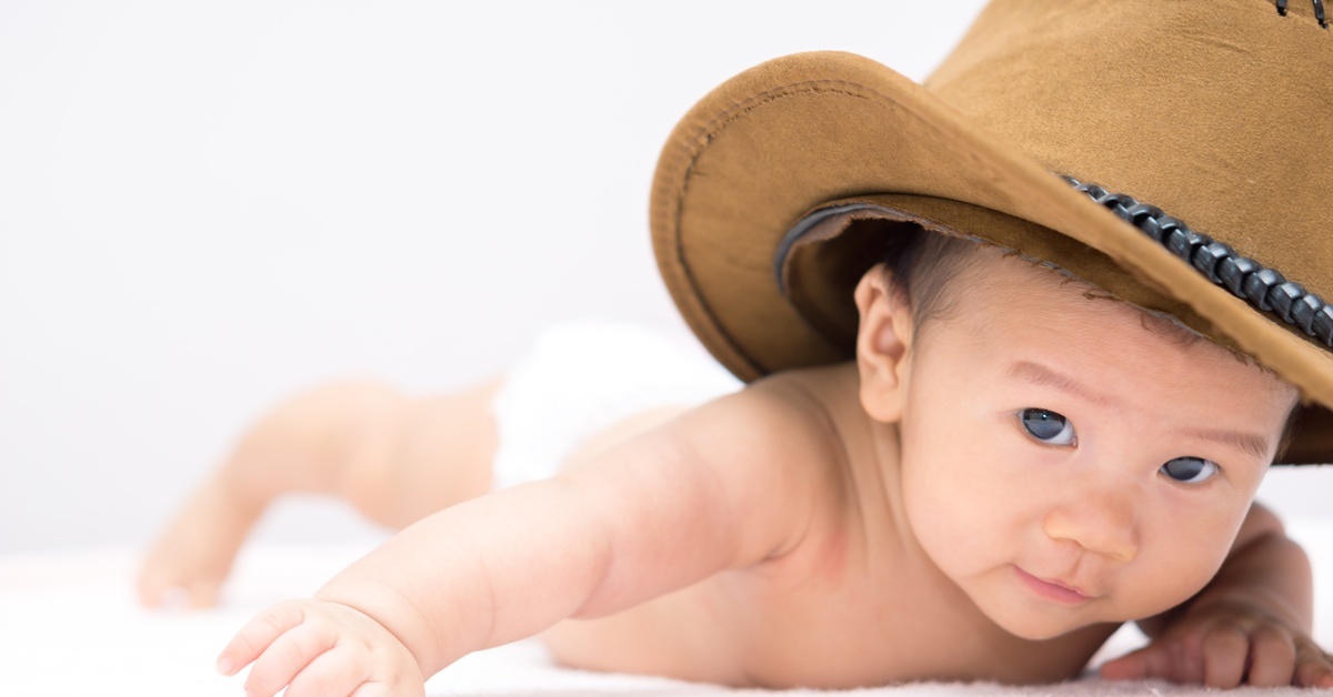 A baby in a diaper and a decorated light tan cowboy hat crawling on the floor while curiously looking around. A baby in a diaper and a decorated light tan cowboy hat crawling on the floor while curiously looking around.