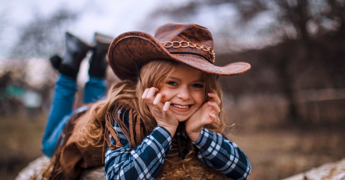 A young girl wearing a large cowboy hat, plaid shirt, jeans, and boots, poses on a hay bale outdoors.