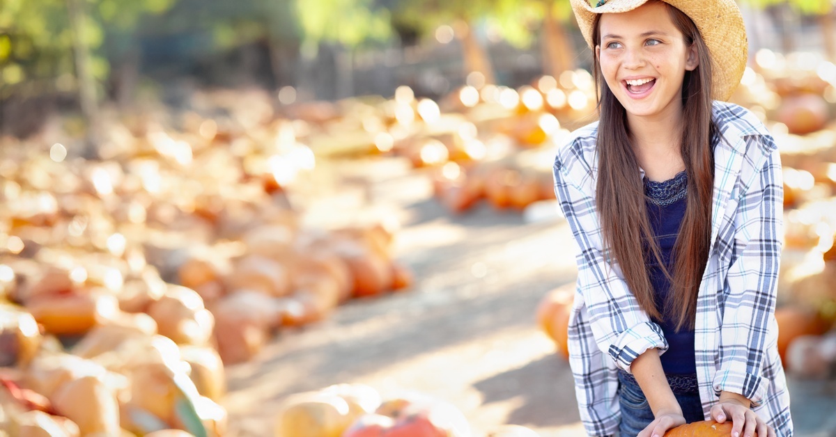 A smiling young girl wearing a straw hat stands in a pumpkin patch, as she holds onto an orange pumpkin.