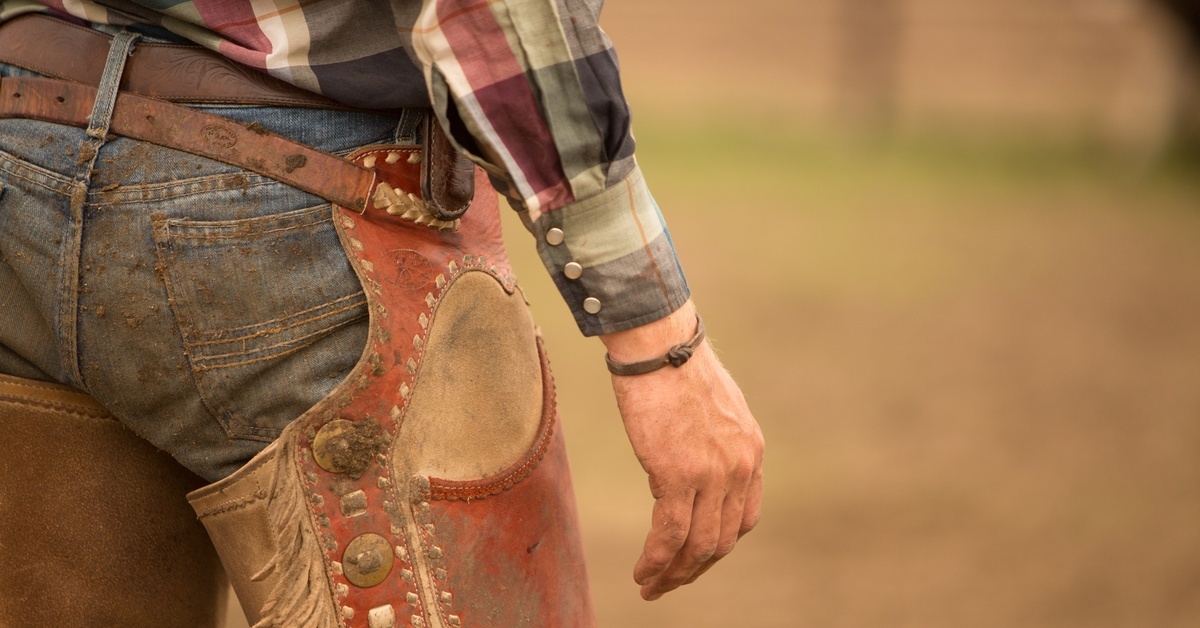 A man wearing western attire, including jeans, leather chaps, a plaid shirt, and a twisted bracelet, stands outside.