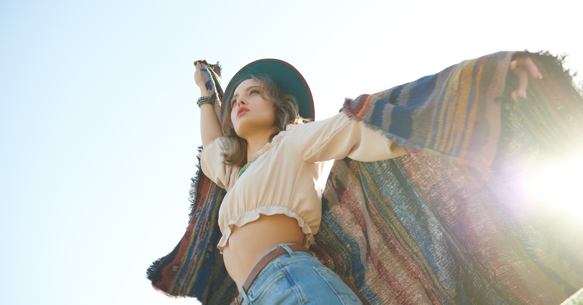 A woman wearing a crop top, jeans, hat, and several accessories twirls with a poncho under a bright, sunny blue sky.