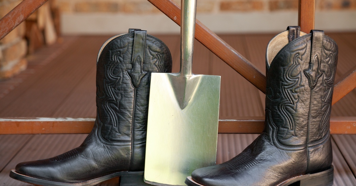 Two black cowboy boots, one on either side of a metal shovel, sitting on a deck in front of a brick wall.