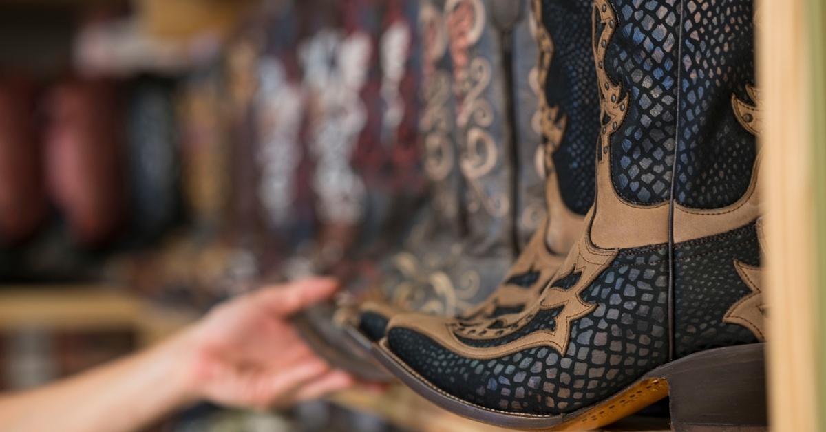 A hand reaching toward a pair of cowboy boots, with a black snakeskin pattern and brown leather accents, on a wooden shelf.