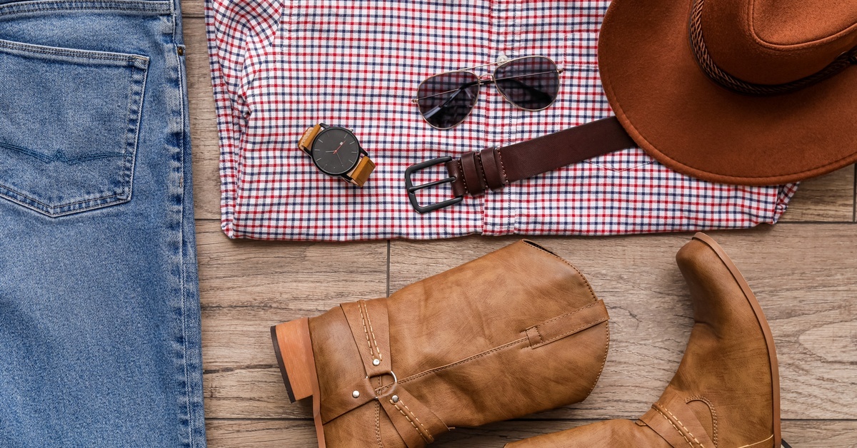 Several items of clothing on a floor including a checkered shirt, jeans, cowboy boots, and accessories like a belt and hat.