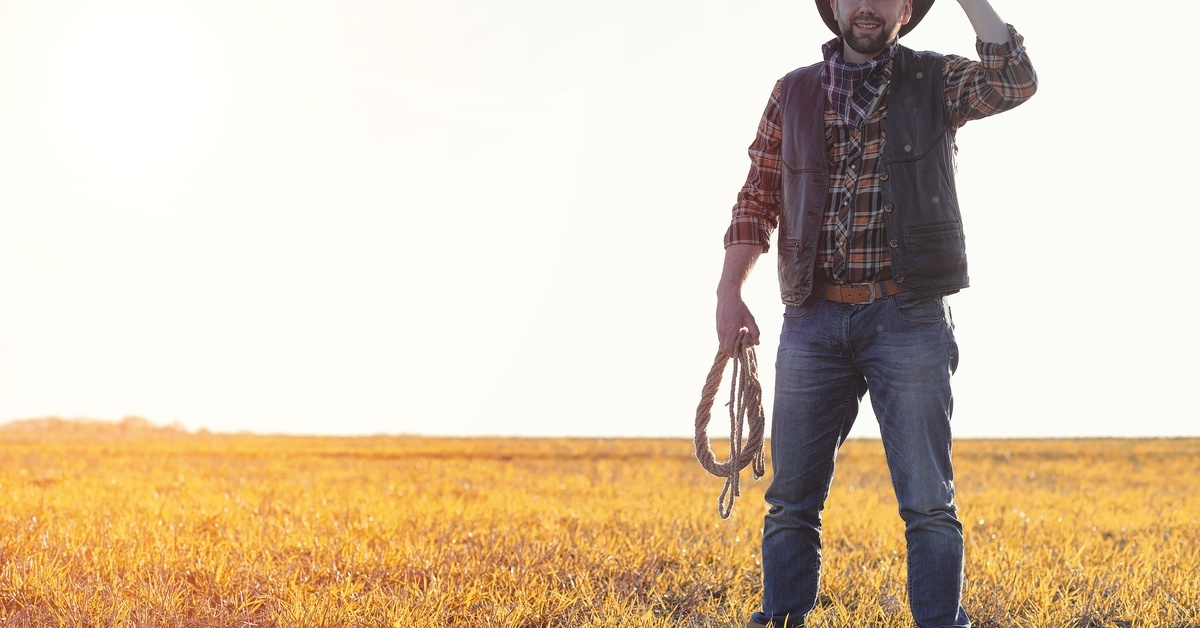 A man wearing a cowboy hat, plaid shirt, vest, and jeans standing in an open grassy field holding a lasso.