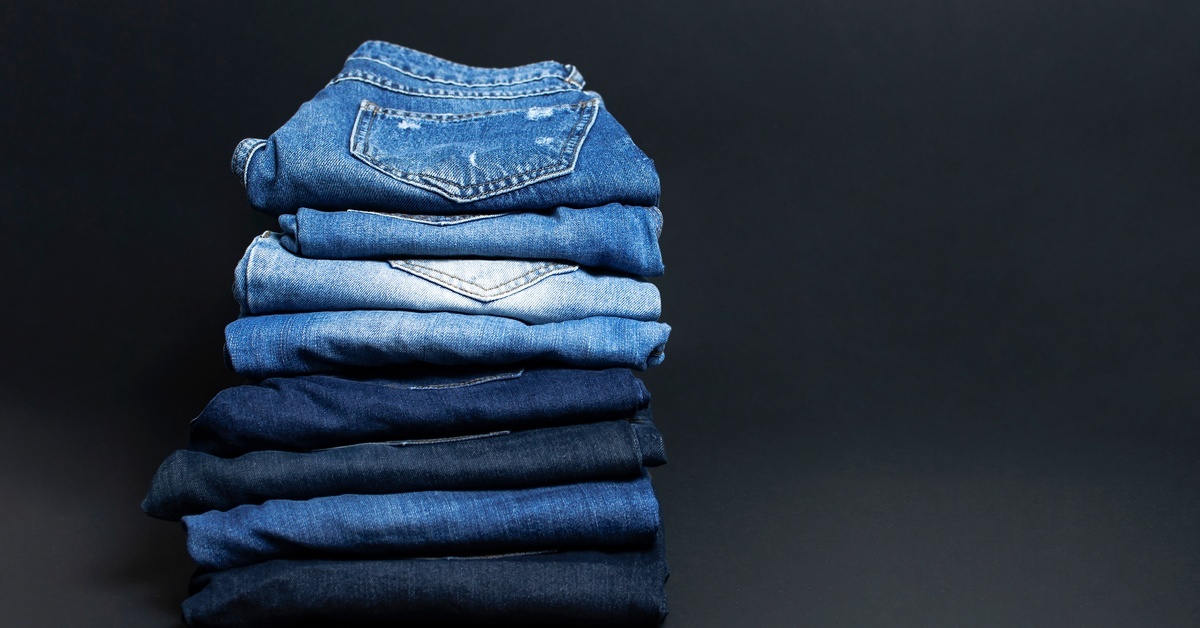 Eight folded pairs of jeans in various colors stacked in a gradient arrangement against a black background.