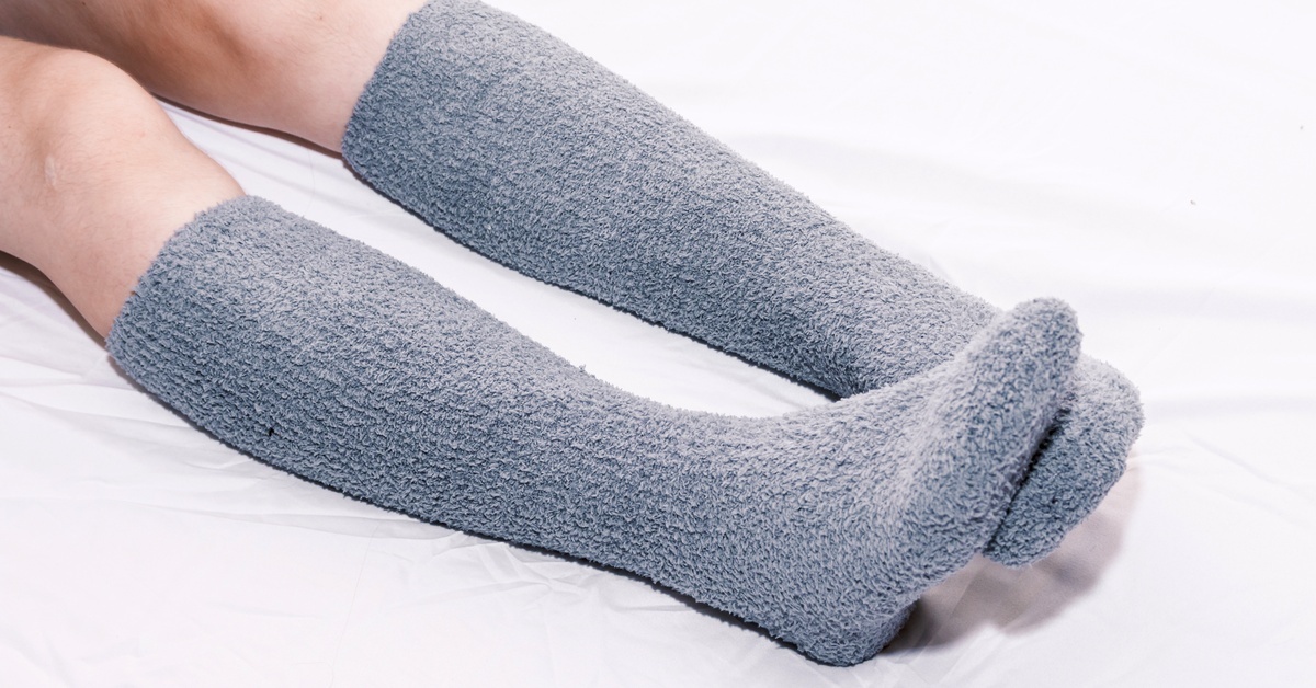 A person relaxing on a sheet-covered mattress, wearing thick, fuzzy gray socks that reach just below their ankles.