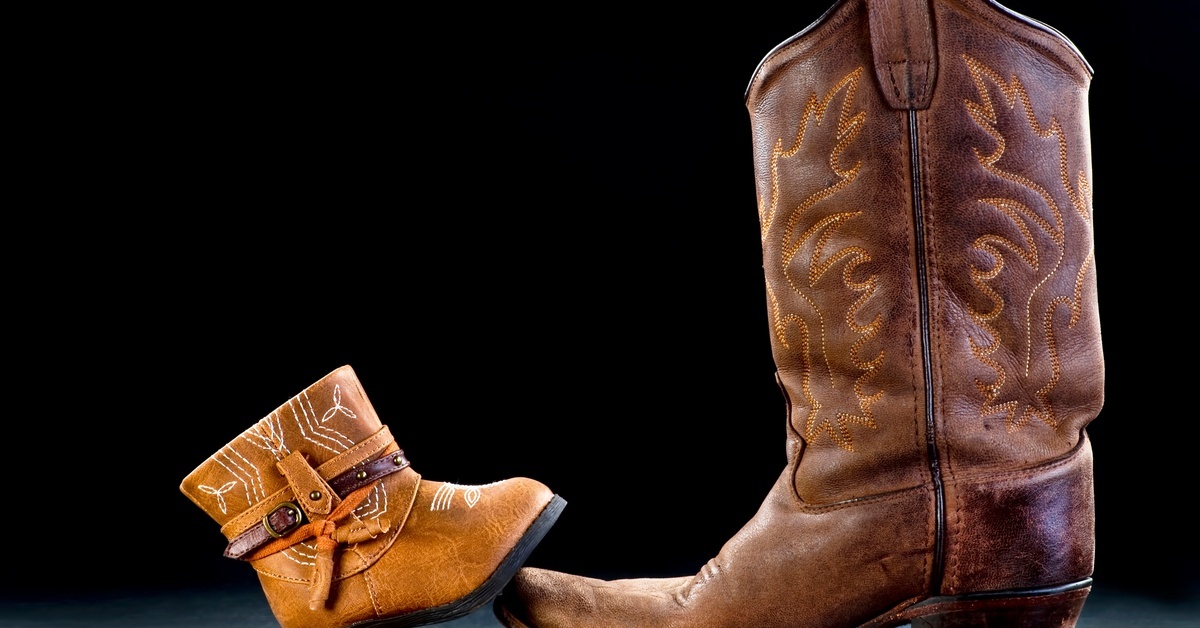 A pair of cowboy boots: one large, adult-sized boot and a smaller child-sized boot stepping onto the larger one's sole.