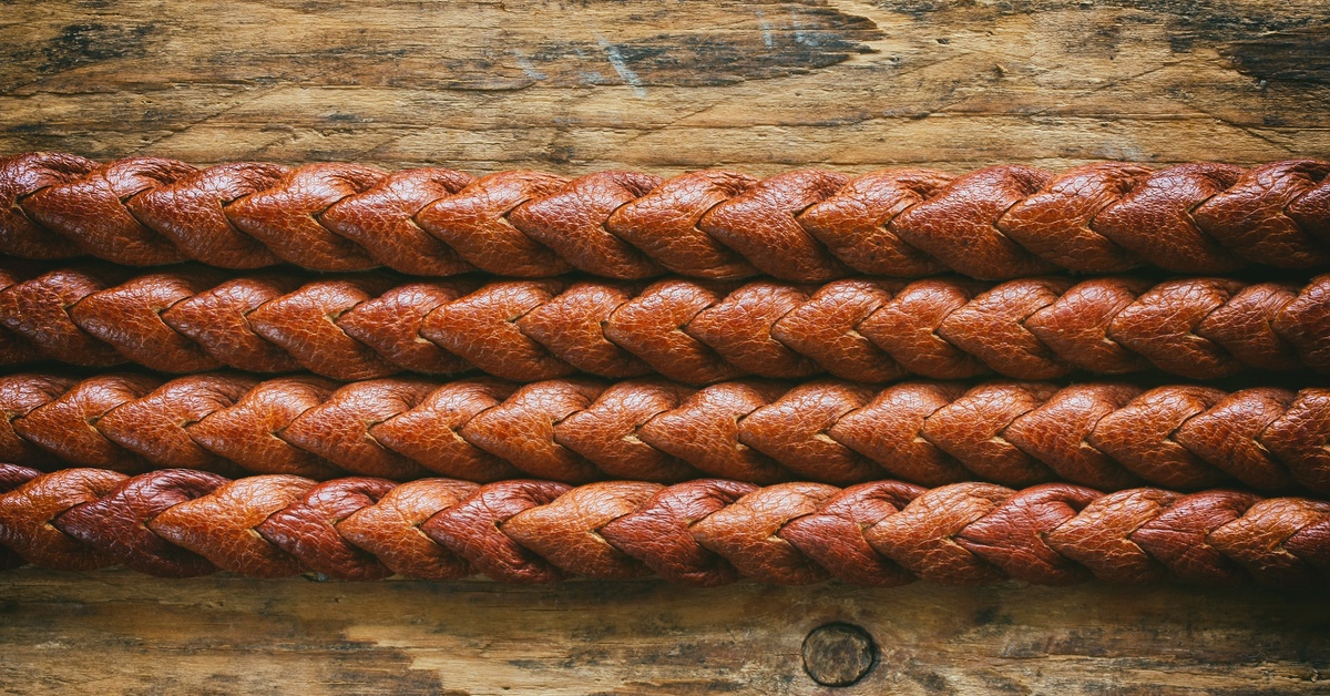 A close-up of the intricate detailing on a braided leather belt laid out on a slightly scratched wooden surface.