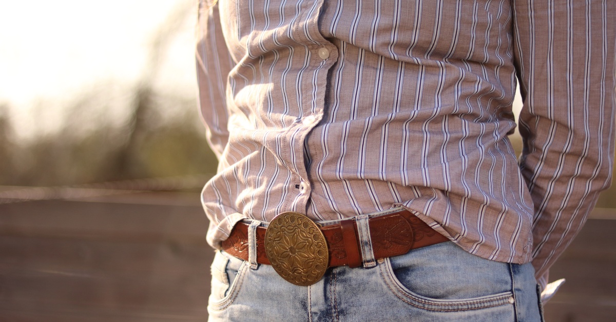 A person in a striped button-down shirt, light-colored jeans, and a western belt leaning against an outdoor wooden fence.