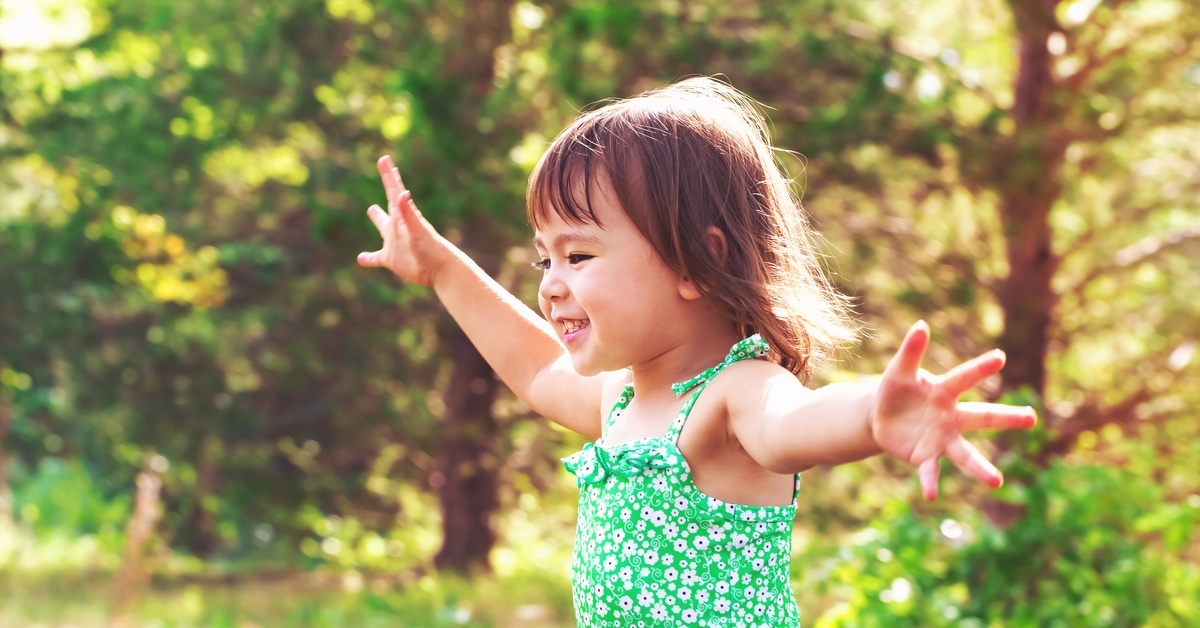 A young smiling girl in a green floral frilly tank top standing outdoors near trees with her arms raised high.