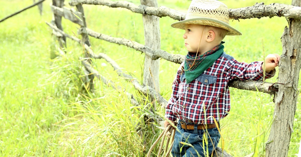 A toddler boy dressed in western gear&mdash;a straw hat, bandana, plaid shirt, and jeans&mdash;leaning against a gnarled wooden fence.