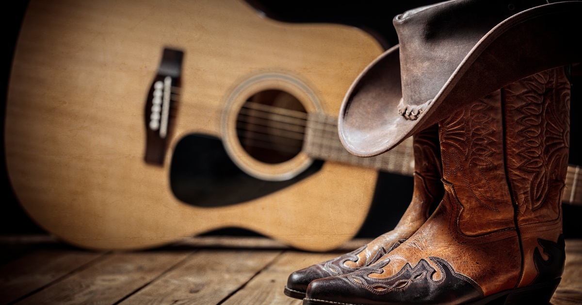 A pair of cowboy boots on a plank floor, with a cowboy hat resting on top. Next to them is a wooden guitar.