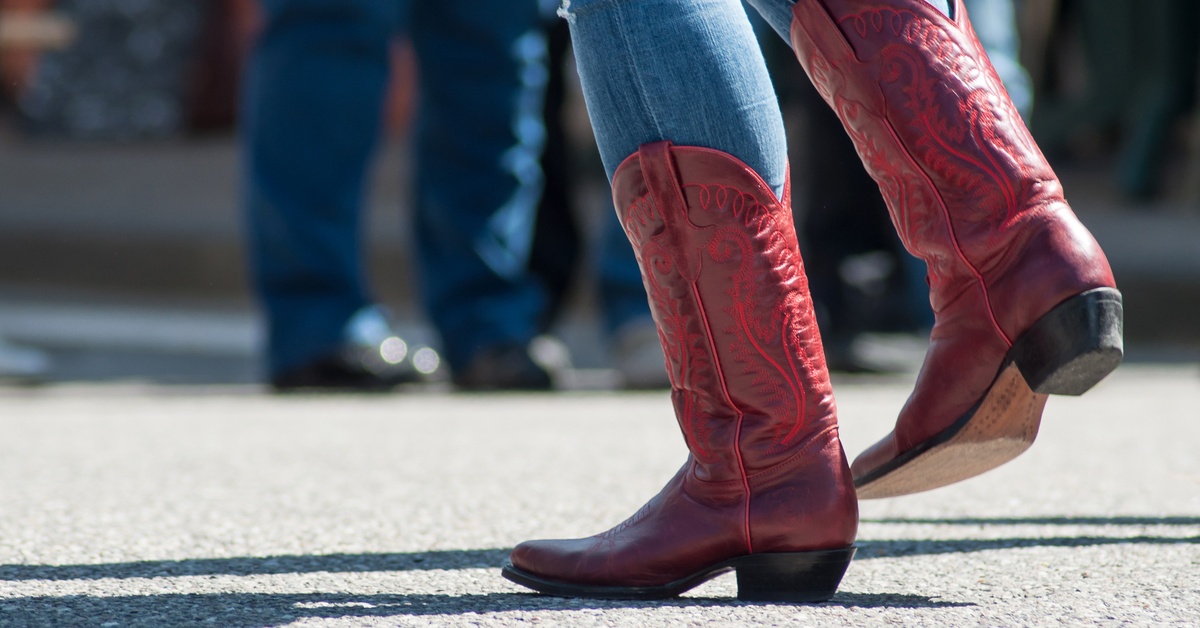 A close-up shot of the lower legs of someone wearing denim jeans and tall red cowboy boots as they walk through a crowd.