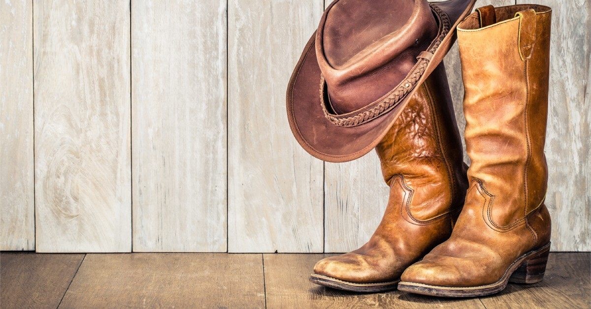 A pair of rounded-toe leather cowboy boots sitting against a rustic wooden wall, topped with a cowboy hat.
