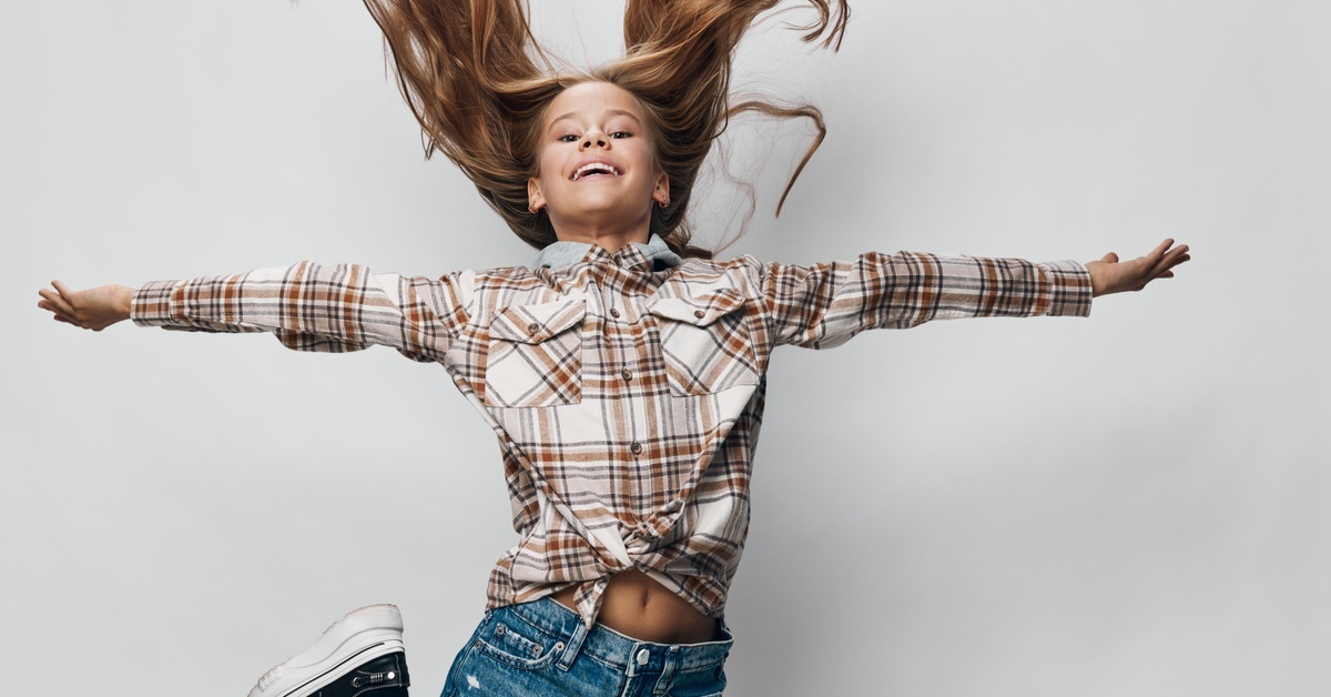 A girl wearing a plaid shirt, denim shorts, and sneakers jumping high into the air with her arms spread out at her sides.