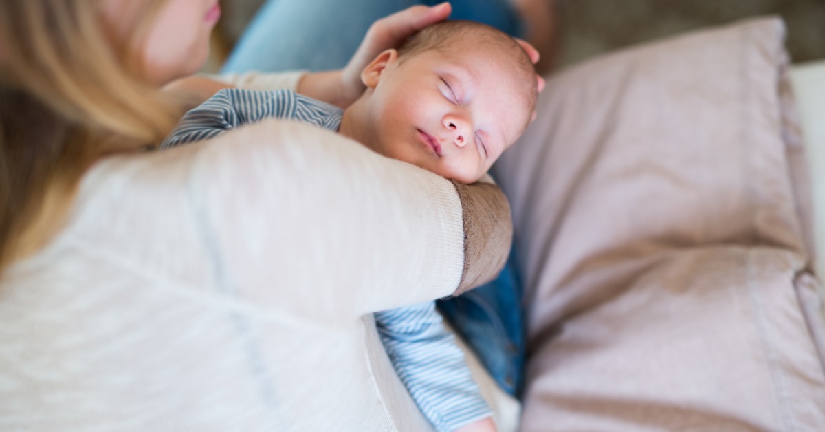 A newborn wearing a simple, striped onesie napping while being cradled by a woman sitting on a couch.