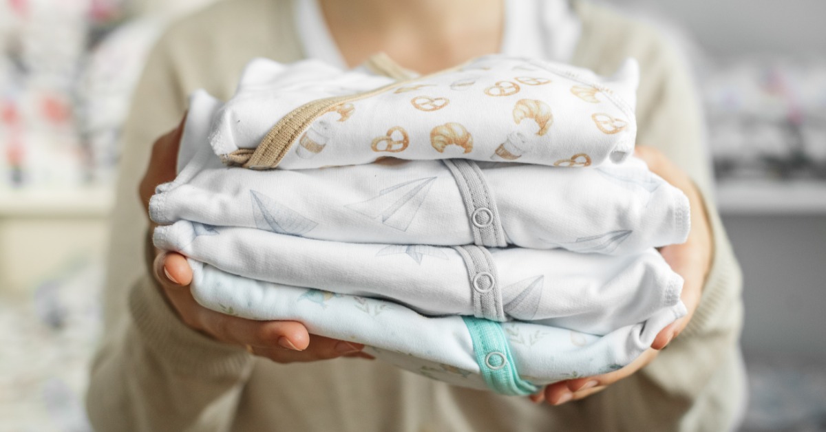 A woman holding a stack of four baby onesies, each a different color and with a different pattern on them.