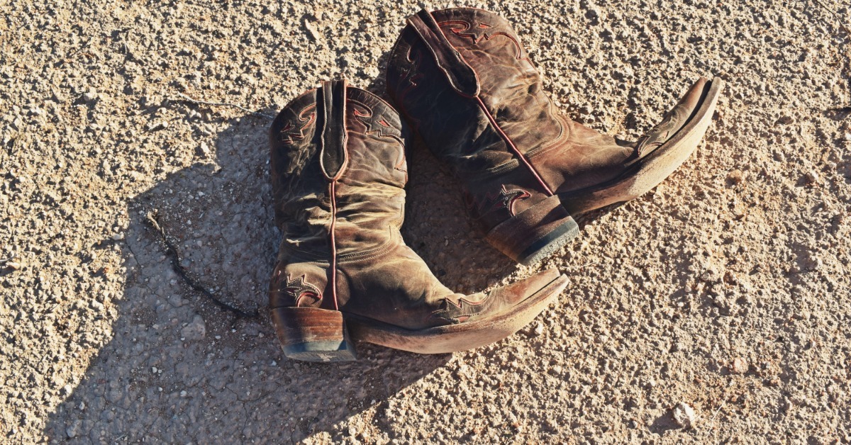 A pair of dusty leather cowboy boots lying side-by-side on rough, gravelly ground, with a large shadow beneath them.