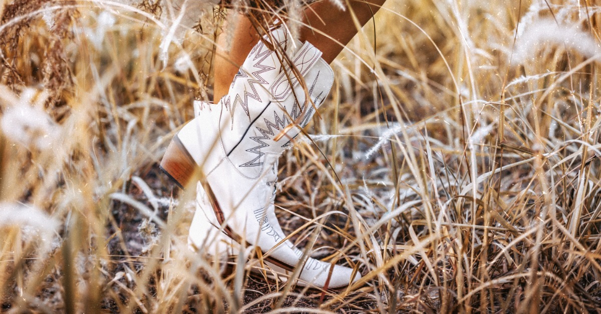 A person wearing white cowboy boots and a woven poncho frolicking through a patch of tall, brown grass.