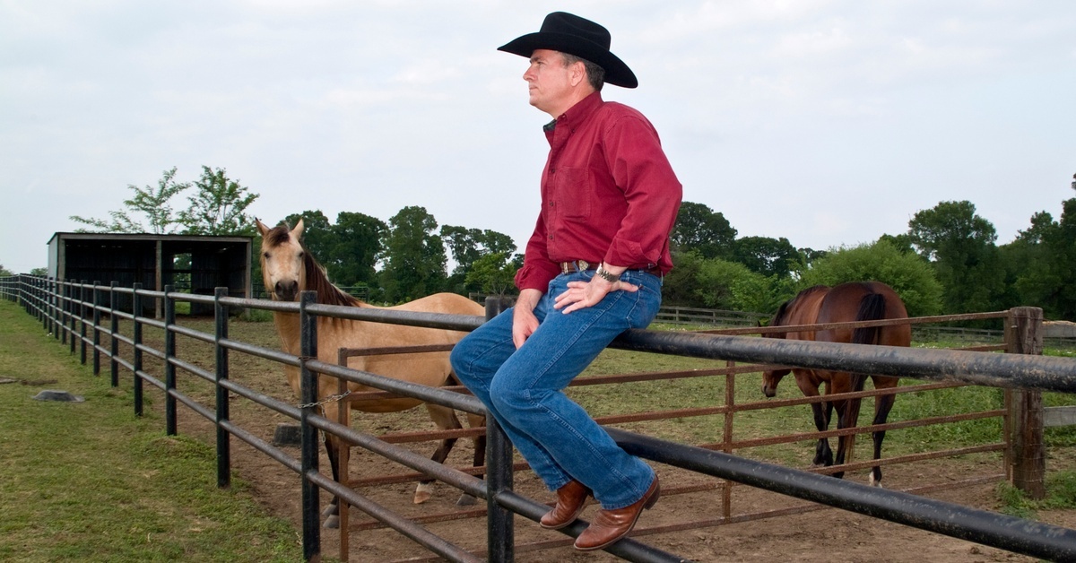 A man wearing a cowboy hat and cowboy boots sitting on a metal fence surrounding a pasture full of horses.