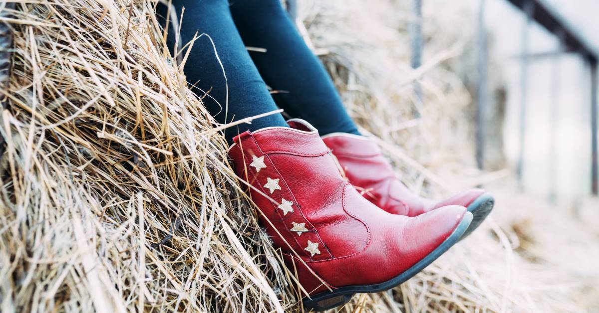 A child wearing jeans and bright red cowboy boots with white stars while sitting on a hay bale outdoors.