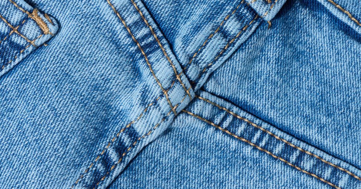 A close-up of blue denim fabric showing reinforced stitching along a seam with detailed texture and thread patterns.