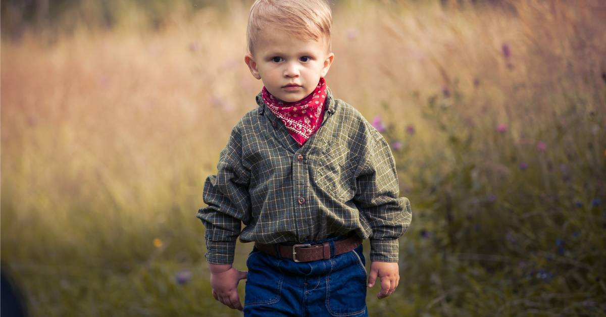 A young boy wearing a western outfit with a hat, shirt, and accessories while standing and looking forward.