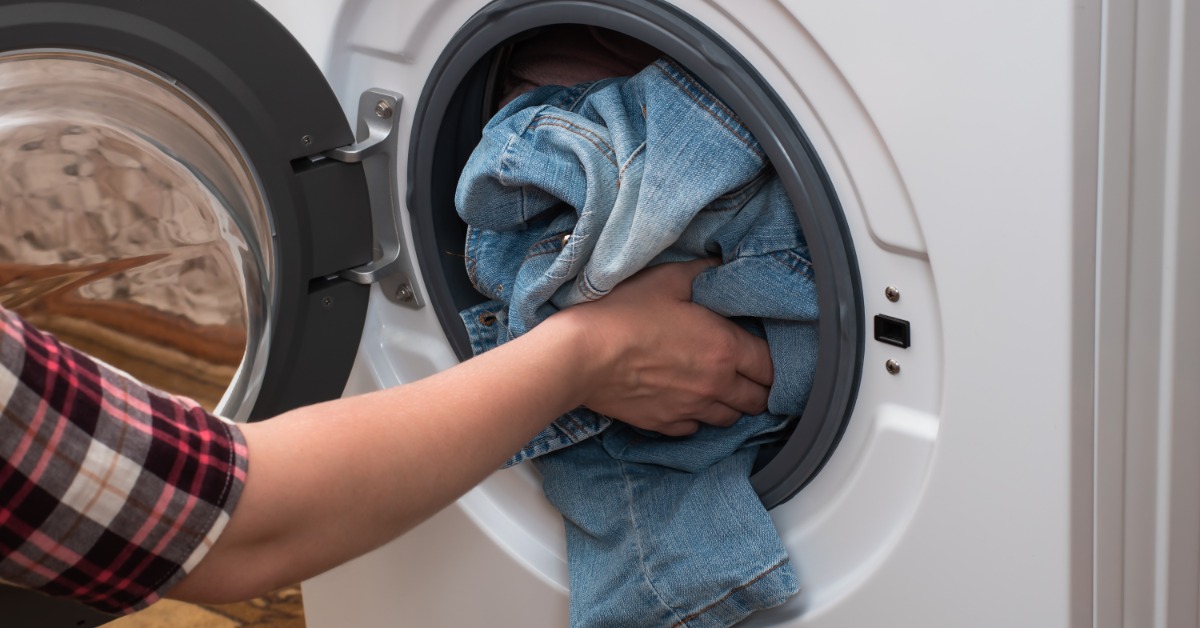 A woman shoving a pair of crumpled blue jeans into a almost-full washing machine while kneeling beside it.