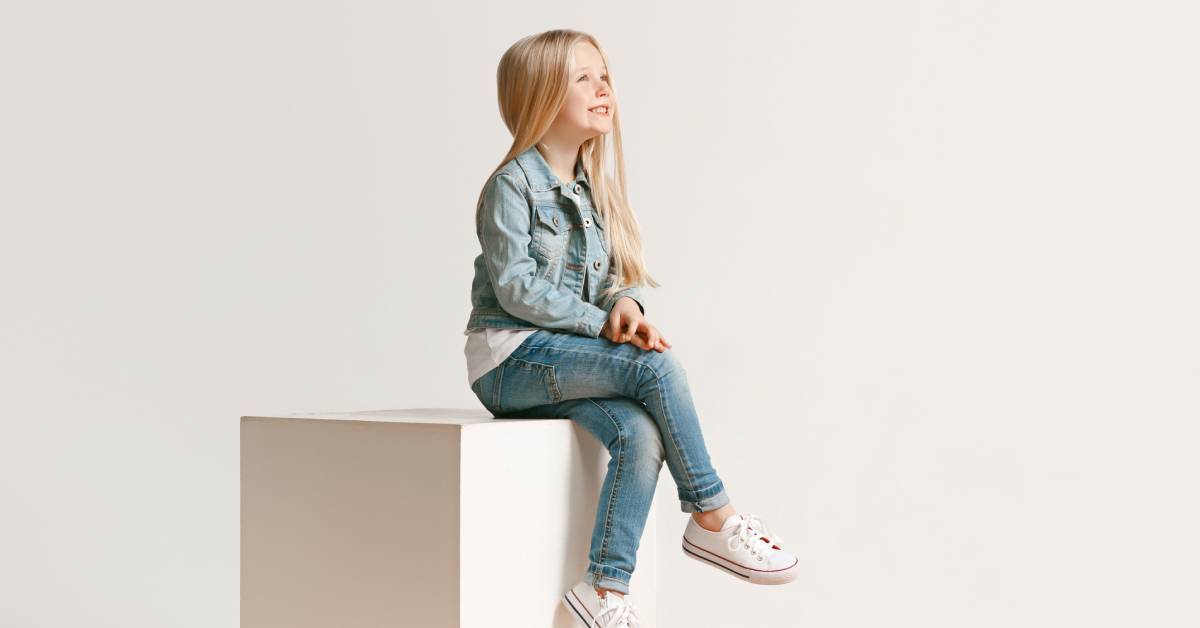 A young girl wearing denim clothing sitting on a cube against a plain background while looking forward with a relaxed posture.