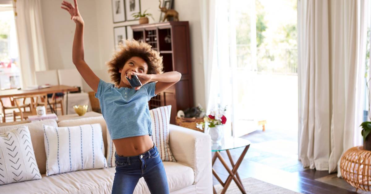 A pre-teen girl standing in a living room while holding a phone like a microphone and moving as she sings along.