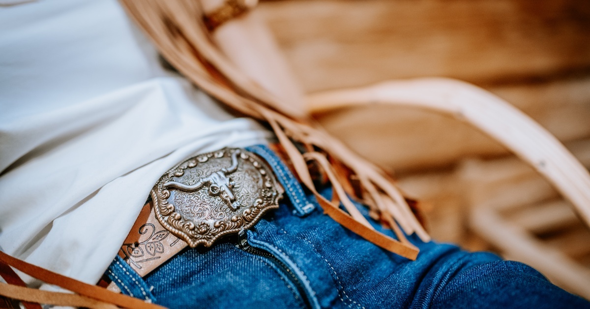 A woman wearing a western outfit with a bull skull belt buckle, denim jeans, white shirt, and fringe vest.