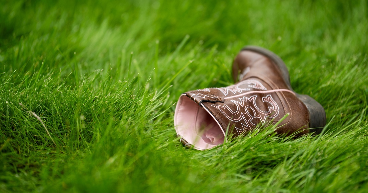 A child's cowboy boot with pink lining lying on its side in lush green grass, shown in a close-up outdoor view.