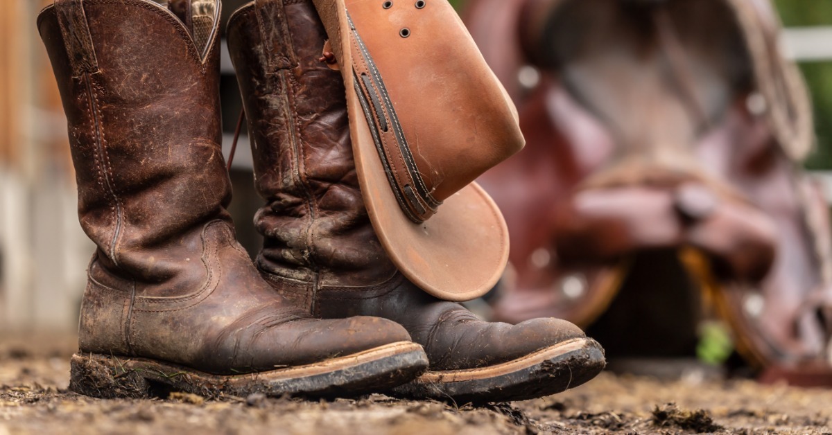 A pair of muddy western boots outside against a wooden exterior wall, with a cowboy hat hanging from one boot.