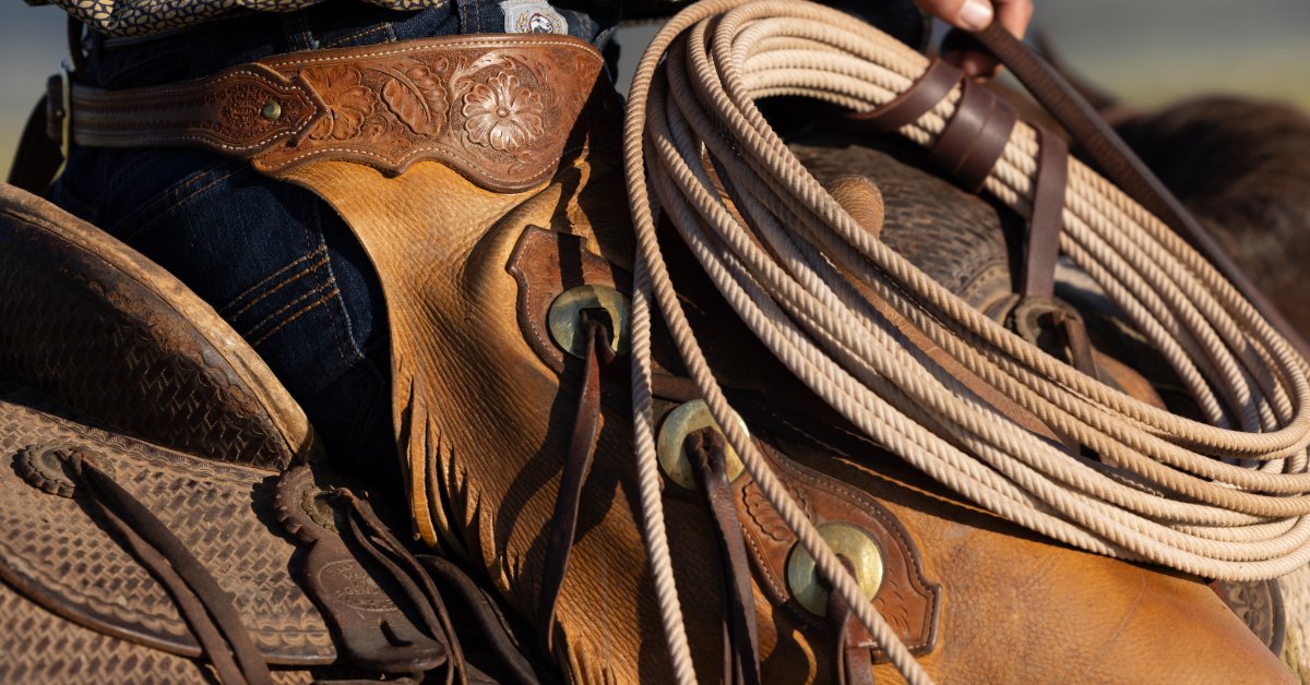 Why Cowgirl Jeans Remain Popular Across Generations A person wearing jeans and leather chaps riding a saddled horse while holding a coiled rope in one hand.
