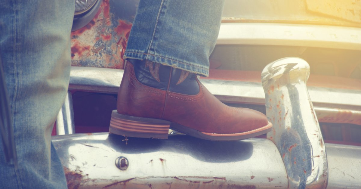 A close-up of a leg in jeans and a cowboy boot perched on the front bumper of an old, rusty antique car.