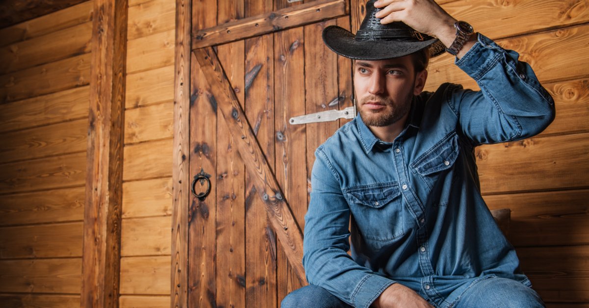 A man with stubble wearing a cowboy hat, denim shirt, jeans, and boots sitting by a wooden wall and door.