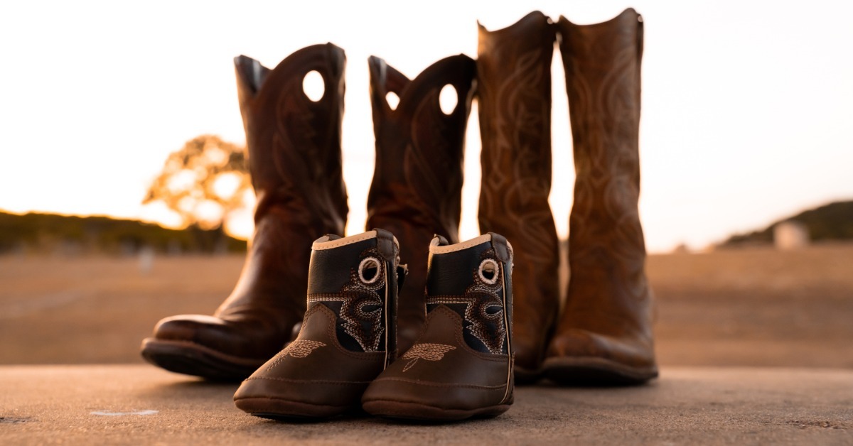 Small brown boots placed in front of taller brown cowboy boots on pavement outdoors at sunset with blurred fields behind.