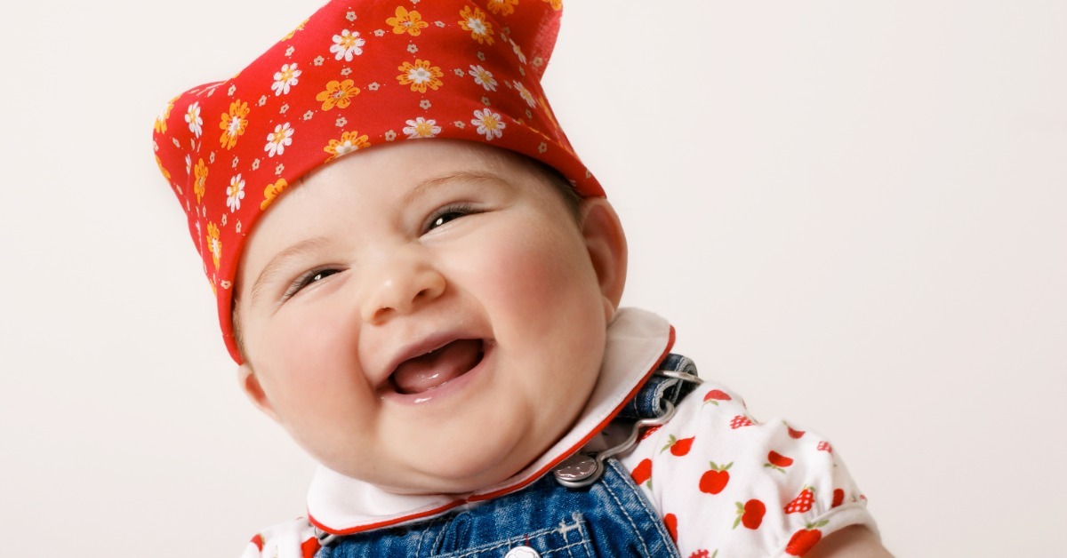 A smiling baby wearing a red floral bandana, apple print shirt, and denim overalls against a plain backdrop.