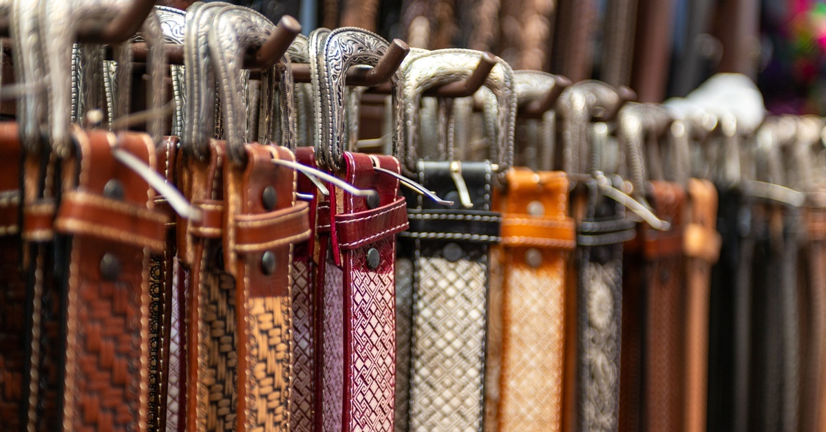 A row of patterned leather belts in assorted colors hanging from hooks with metal buckles facing forward. A row of patterned leather belts in assorted colors hanging from hooks with metal buckles facing forward.