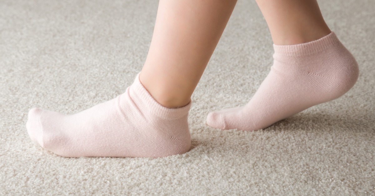 A close-up side view of a child's lower legs wearing pale pink socks while walking on a beige carpet.