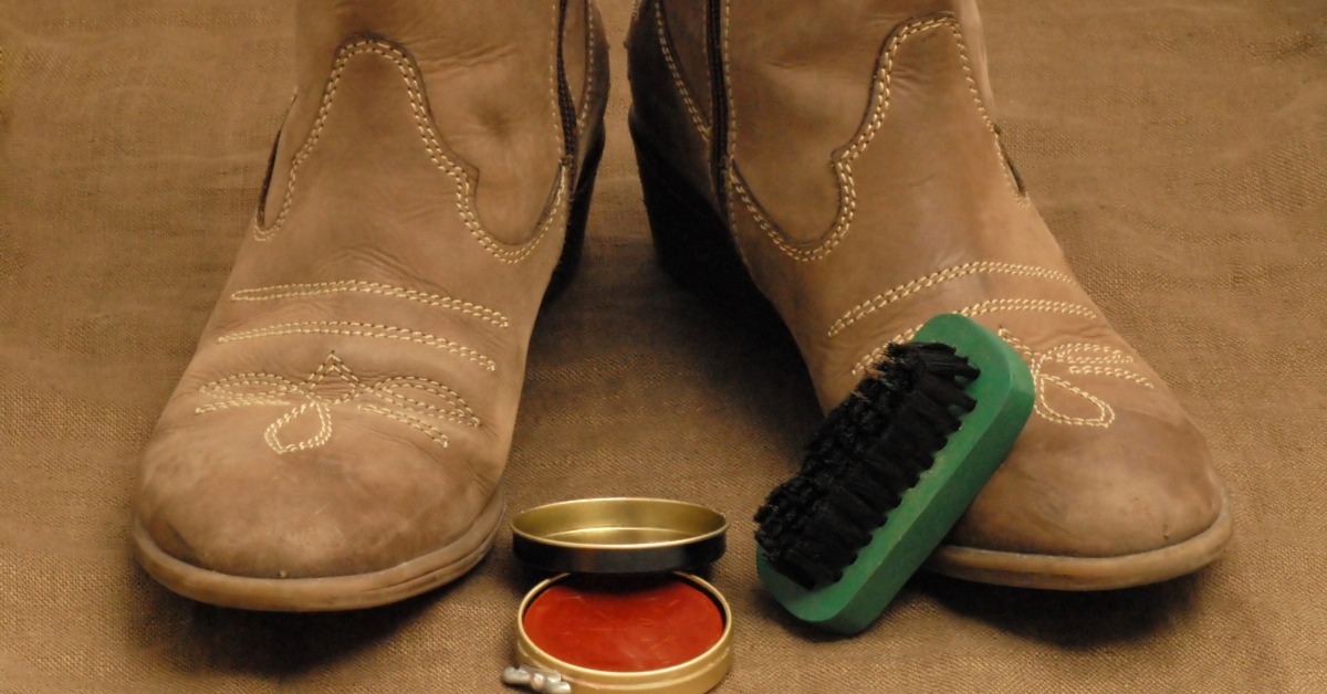 A pair of tan boots on brown fabric with an open polish tin and green brush placed in front of them.