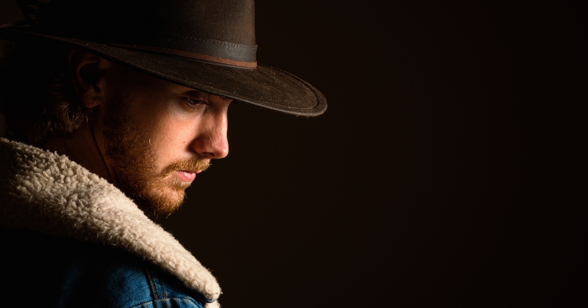 A bearded man wearing a dark cowboy hat and shearling jacket against a dark, plain background with side lighting on his face.