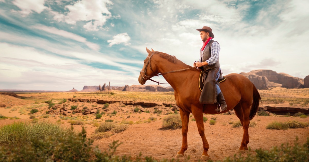 A man wearing a cowboy hat and red neckerchief riding a brown horse through a desert with mesas and clouds.