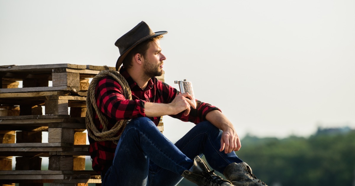 A man in a cowboy hat and red plaid shirt sitting beside wooden pallets while holding a metal flask outdoors.