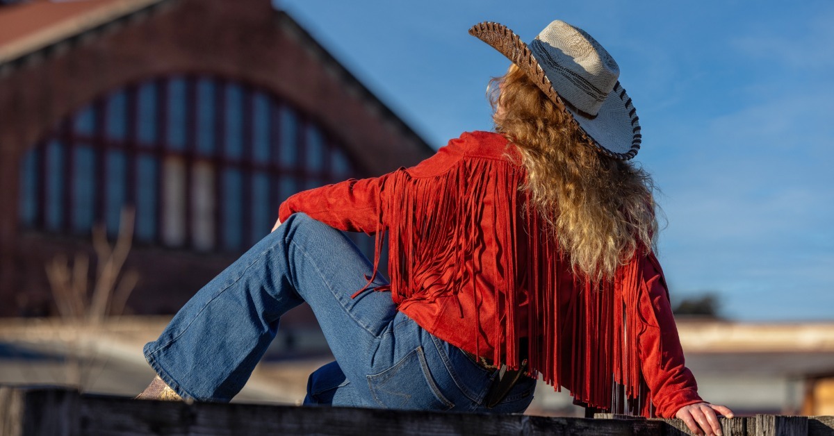 A woman wearing a cowboy hat, red fringe jacket, and blue jeans sitting on a wooden fence near a brick building.